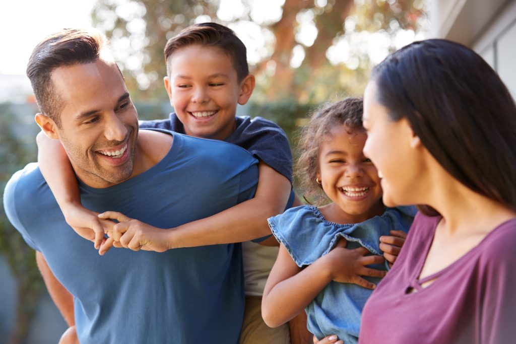 Smiling Family With Parents Giving Children Piggyback Rides In Garden At Home